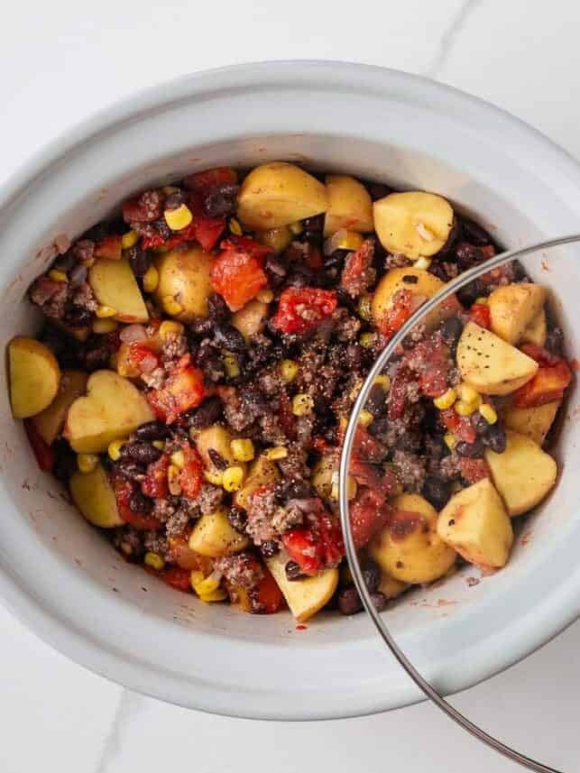 Placing the lid on a slow cooker filled with ground beef, potatoes, beans, corn and tomatoes.