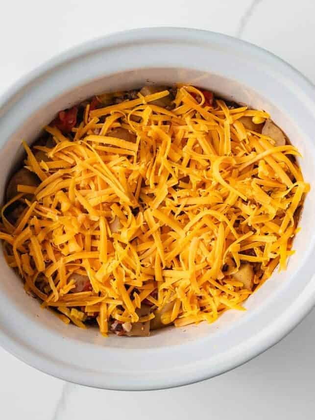 Shredded cheddar cheese sprinkled over cowboy casserole ingredients in a crockpot.