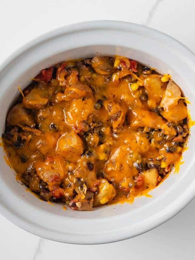 Cooked cowboy casserole topped with melted cheddar cheese in a crockpot.