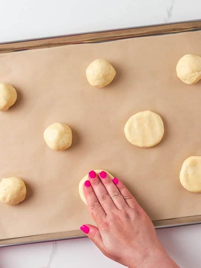 Sugar cookie dough balls being flattened by hand on a parchment-lined baking sheet.