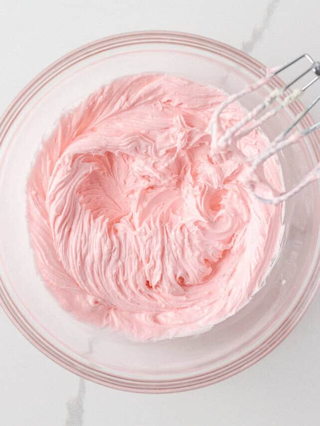 Crumble signature pink buttercream frosting with almond extract whipped in a glass bowl with an electric mixer.