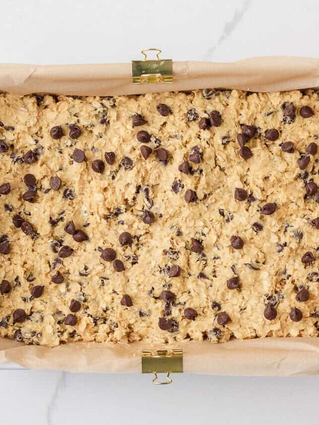 Oatmeal chocolate chip cookie bar dough pressed evenly into a parchment-lined baking dish.