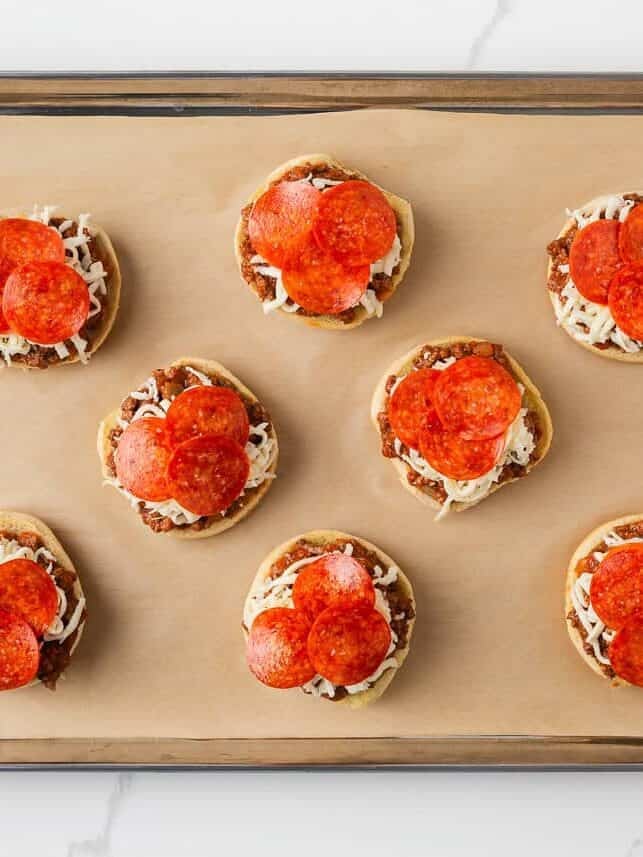 Open-faced pizza burgers assembled on a lined baking sheet before baking with cheese and pepperoni topping.