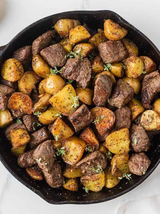 Cast iron skillet filled with garlic butter steak bites and seasoned golden potatoes topped with fresh thyme.