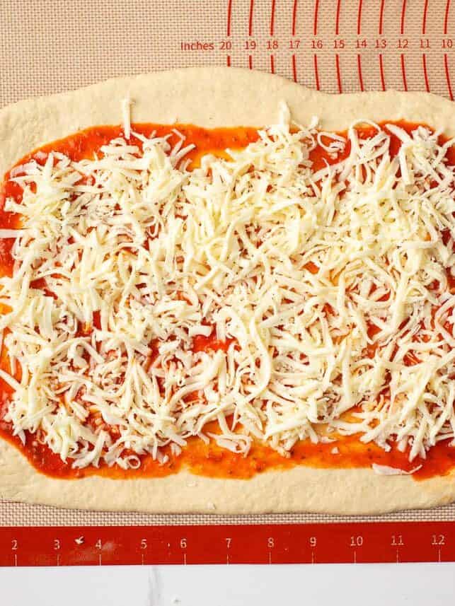 Shredded mozzarella cheese layered over pizza sauce on rolled pizza dough.