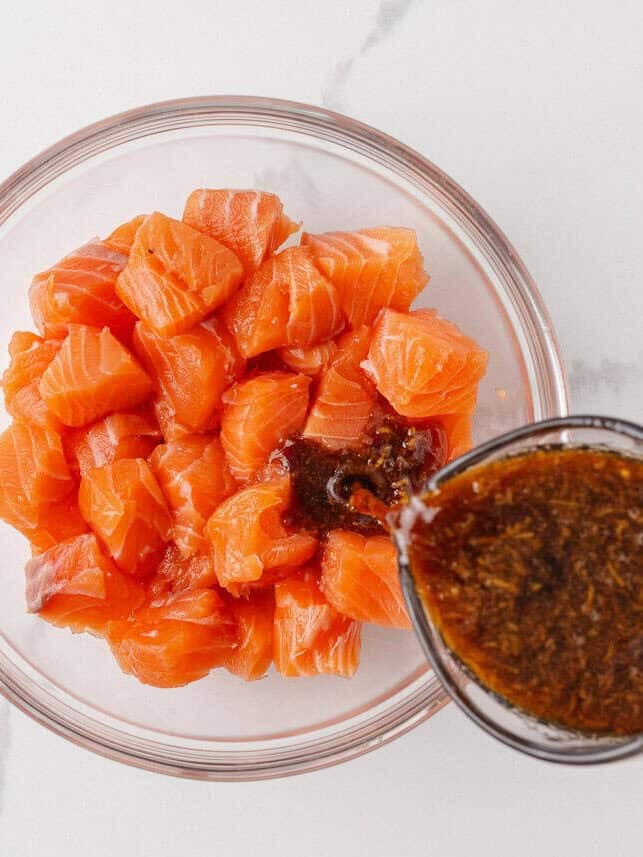 Homemade teriyaki marinade being poured over cubed salmon in a glass bowl.