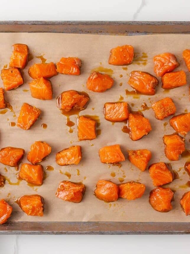 Broiled teriyaki salmon bites on a parchment-lined baking sheet with wooden kitchen tongs.