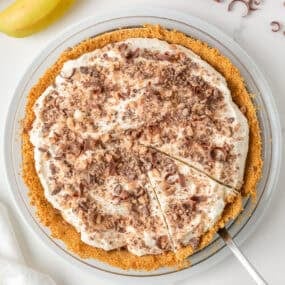 Overhead view of banoffee pie topped with toffee bits and chocolate curls with one slice cut.
