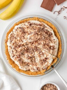 Overhead view of banoffee pie topped with toffee bits and chocolate curls with one slice cut.