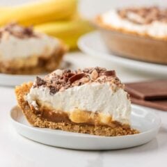 Banoffee pie slice on a white plate with bananas, chocolate and the remaining pie in the background.