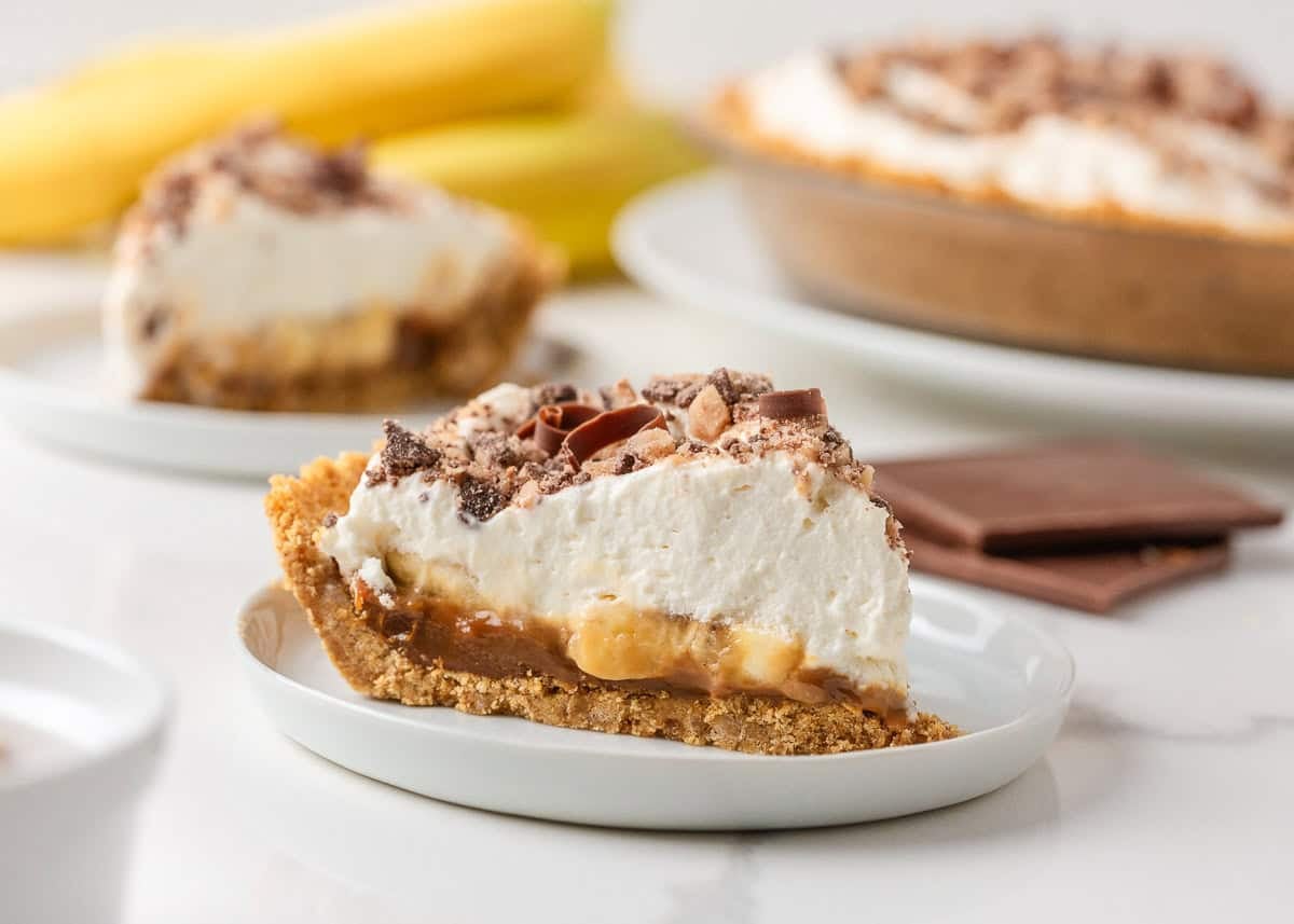 Banoffee pie slice on a white plate with bananas, chocolate and the remaining pie in the background.