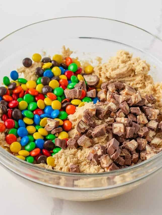 Candy cookie dough topped with colorful M&M’s and chopped chocolate candy bars in a glass mixing bowl.