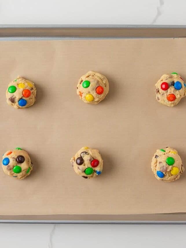 Scoops of candy cookie dough with M&M’s and candy pieces arranged on a parchment-lined baking sheet.