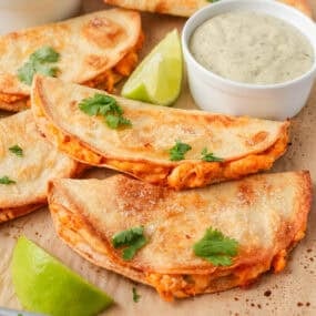 Golden crispy chicken tacos on a baking sheet with salsa, creamy dipping sauce and fresh lime.