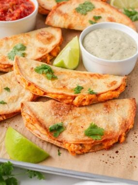 Golden crispy chicken tacos on a baking sheet with salsa, creamy dipping sauce and fresh lime.
