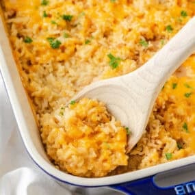 Wooden spoon scooping cheesy chicken and rice casserole with parsley from a baking dish.