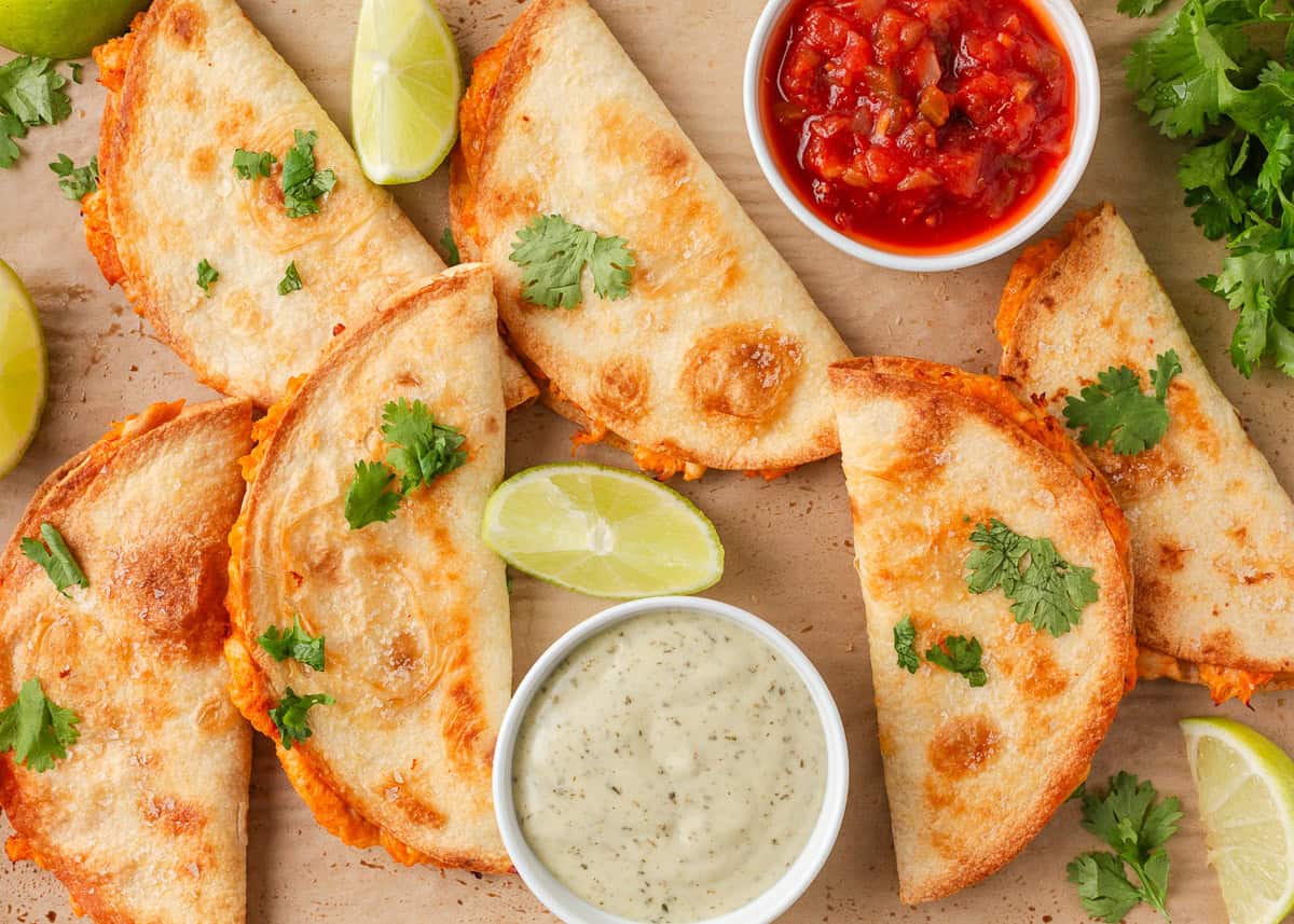 Overhead view of crispy chicken tacos served with salsa, creamy sauce, cilantro and lime wedges.