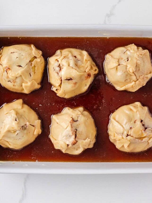 Unbaked apple dumplings wrapped in dough and sitting in brown sugar sauce inside a white baking dish.