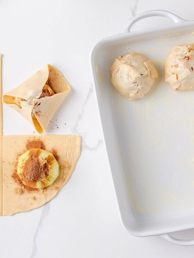 Partially assembled apple dumplings being wrapped in dough and placed into a buttered baking dish.