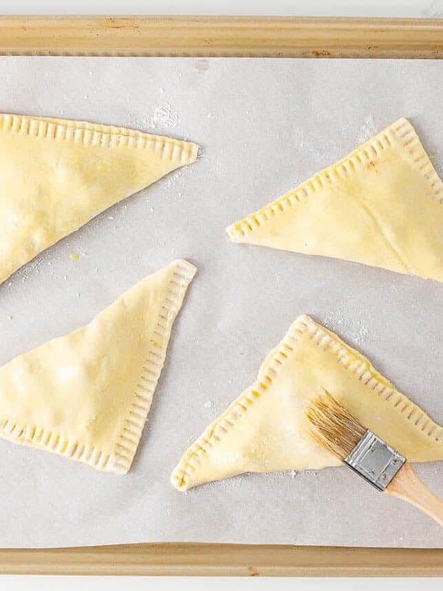 Unbaked apple turnovers sealed with a fork and brushed with egg wash on a baking sheet.