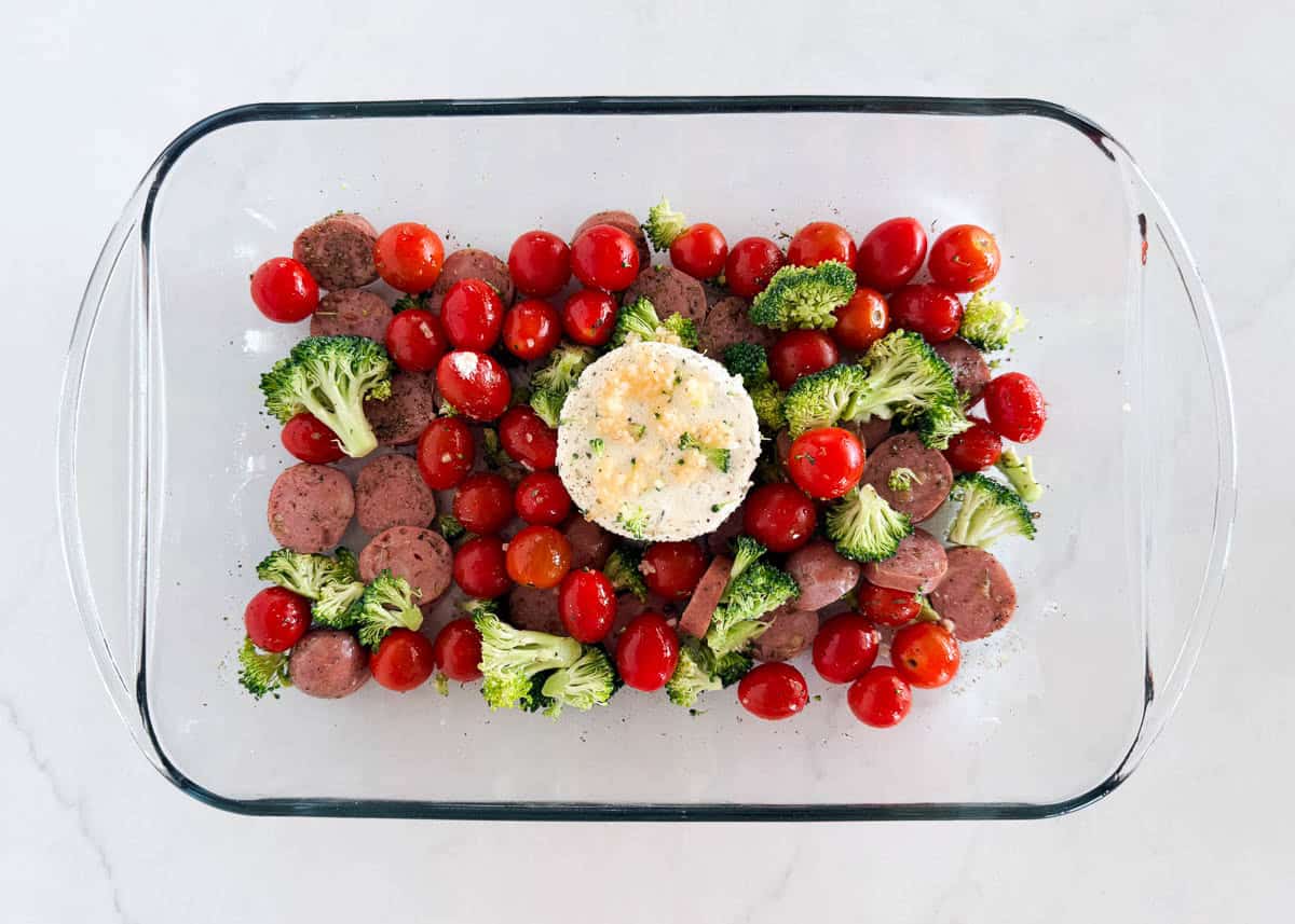 Boursin cheese in a baking dish surrounded by sausage, broccoli and cherry tomatoes.