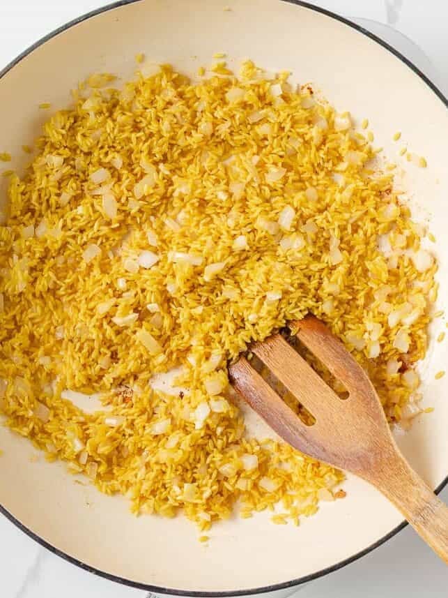 Toasted orzo and onions being stirred with a wooden spoon.