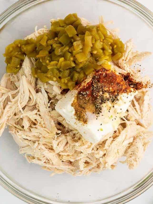 Shredded chicken, cream cheese, diced green chiles and spices in a glass mixing bowl.