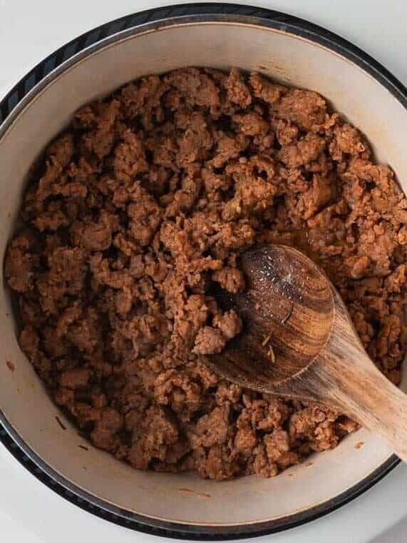 Ground Italian sausage browning in a Dutch oven with a wooden spoon.