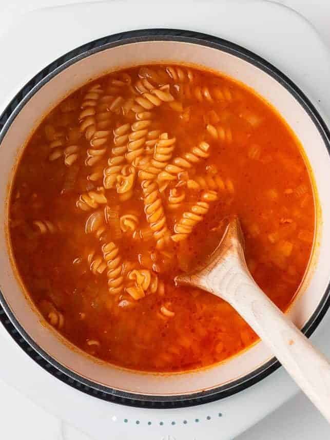 Step showing pasta simmering in tomato broth in a white pot on the stove.