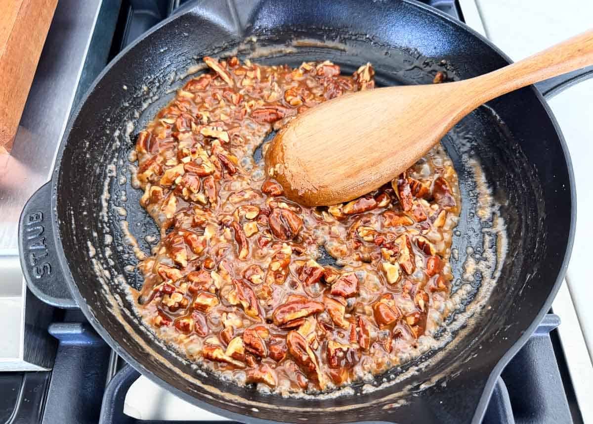 Pecan topping bubbling in a skillet with chopped pecans and a wooden spoon.