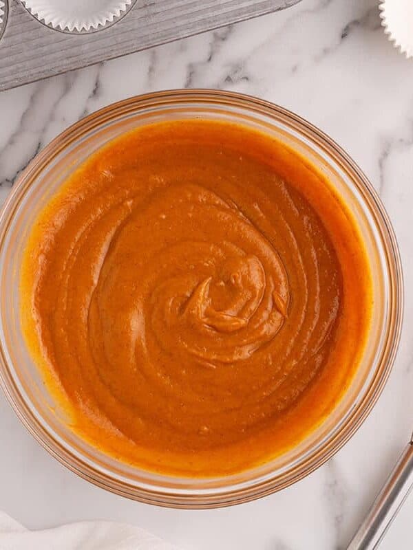 Smooth pumpkin spice cupcake batter in a glass bowl beside a whisk and cupcake liners.
