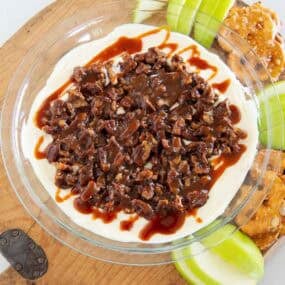 Pecan pie dip topped with chopped pecans and caramel, surrounded by apple slices and pretzels.
