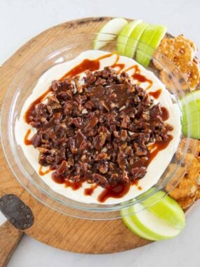 Pecan pie dip topped with chopped pecans and caramel, surrounded by apple slices and pretzels.