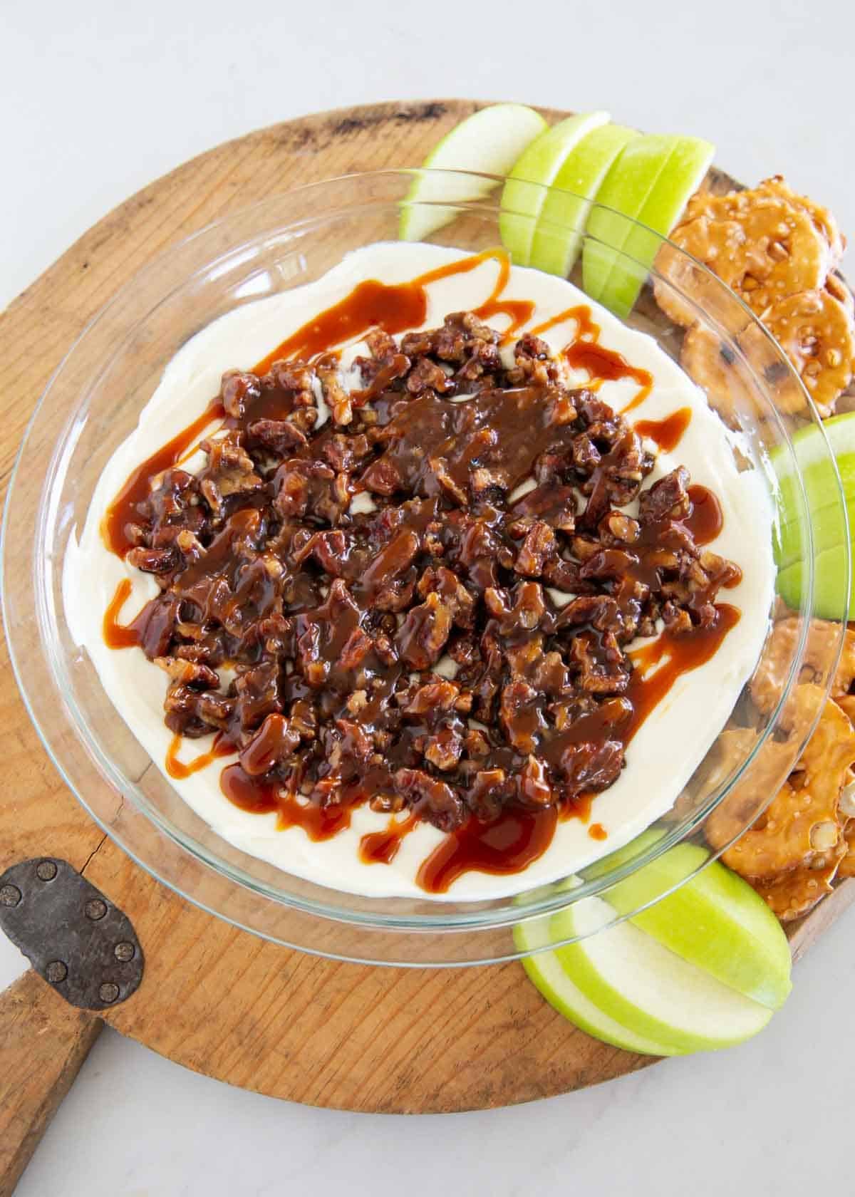 Pecan pie dip topped with chopped pecans and caramel, surrounded by apple slices and pretzels.