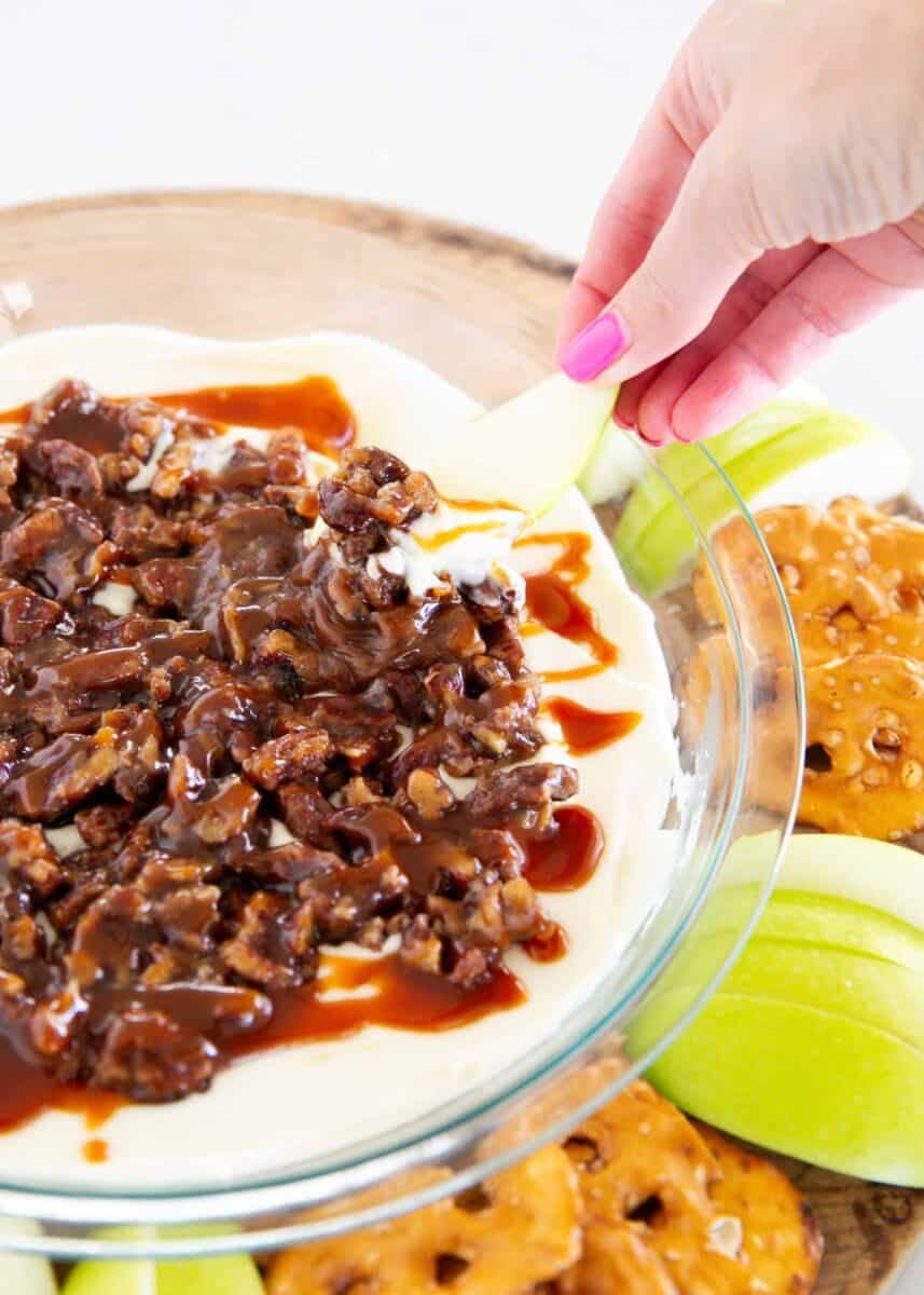 A hand dips an apple slice into pecan pie dip topped with pecans and caramel.