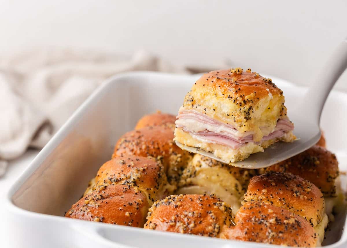 Ham and Swiss cheese slider on a spatula above a pan of Hawaiian roll sliders with poppy seed topping.