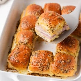 Baked ham and cheese sliders in a casserole dish with one Hawaiian roll slider being served.