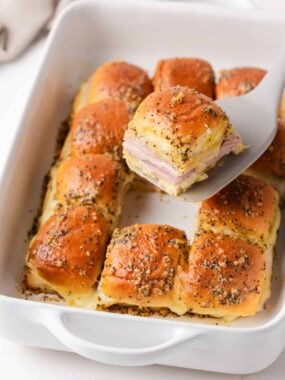 Baked ham and cheese sliders in a casserole dish with one Hawaiian roll slider being served.