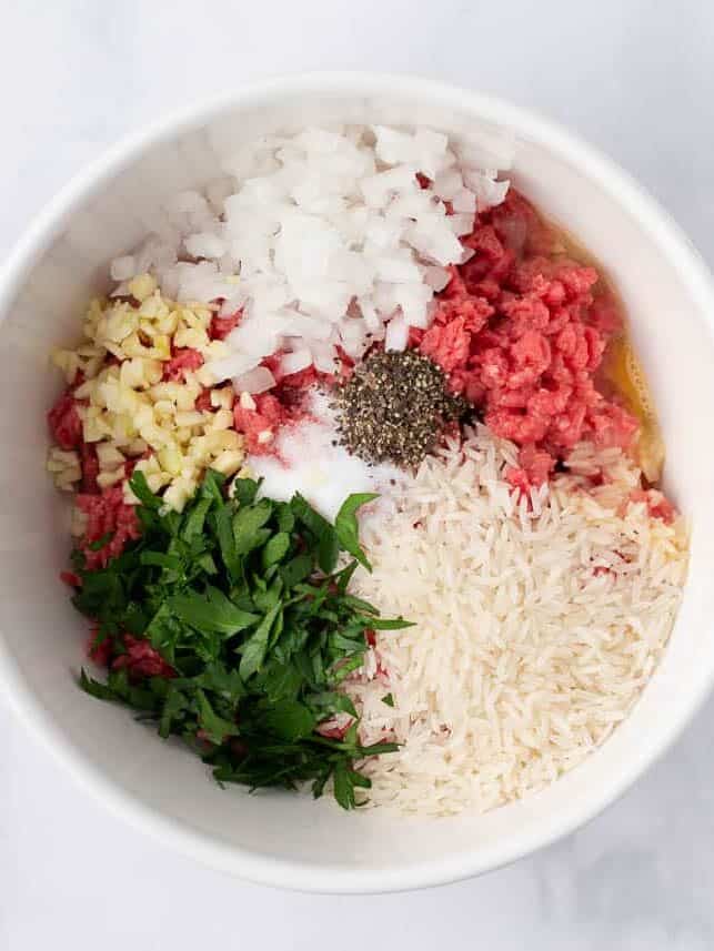 Ground beef, uncooked rice, egg, onion, garlic, parsley, salt, and pepper in a bowl, ready to mix.
