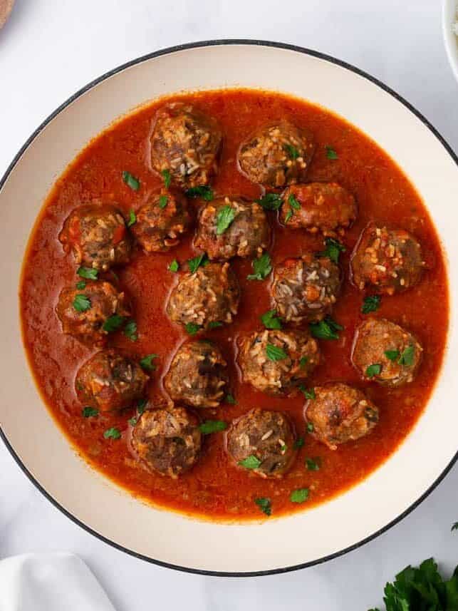 Porcupine meatballs simmering in tomato sauce in a skillet, with rice and parsley nearby.