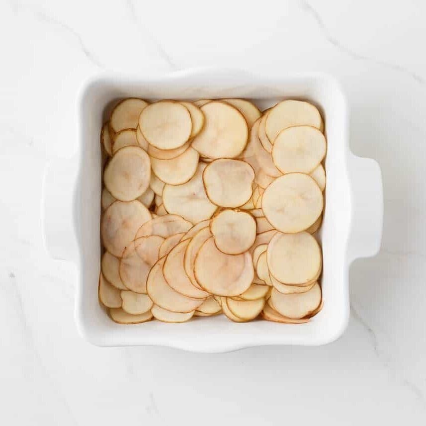 Thinly sliced potatoes arranged in overlapping layers in a white baking dish for homemade potatoes au gratin.