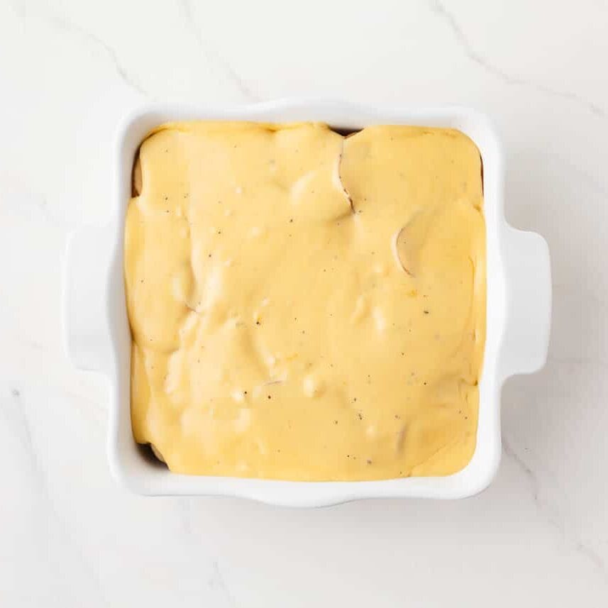Unbaked potatoes au gratin covered in a creamy cheddar cheese sauce in a white casserole dish.
