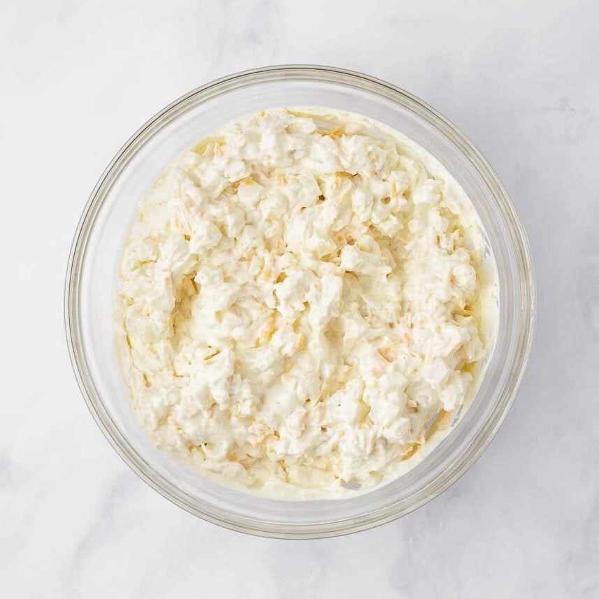 Diced hash browns and shredded cheddar folded into the creamy sauce in a glass bowl, ready to transfer to the baking dish.