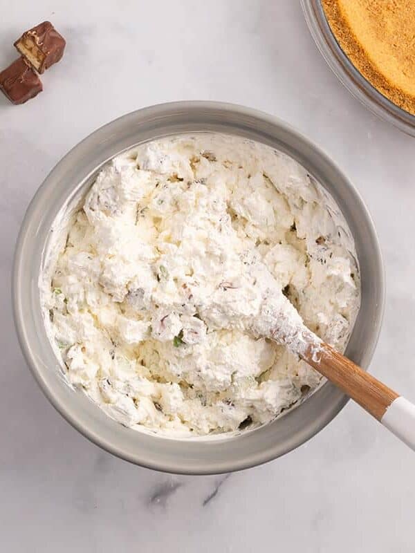 Creamy Snickers apple pie filling mixed in a bowl with chopped apples and candy pieces, ready to add to the graham cracker crust.
